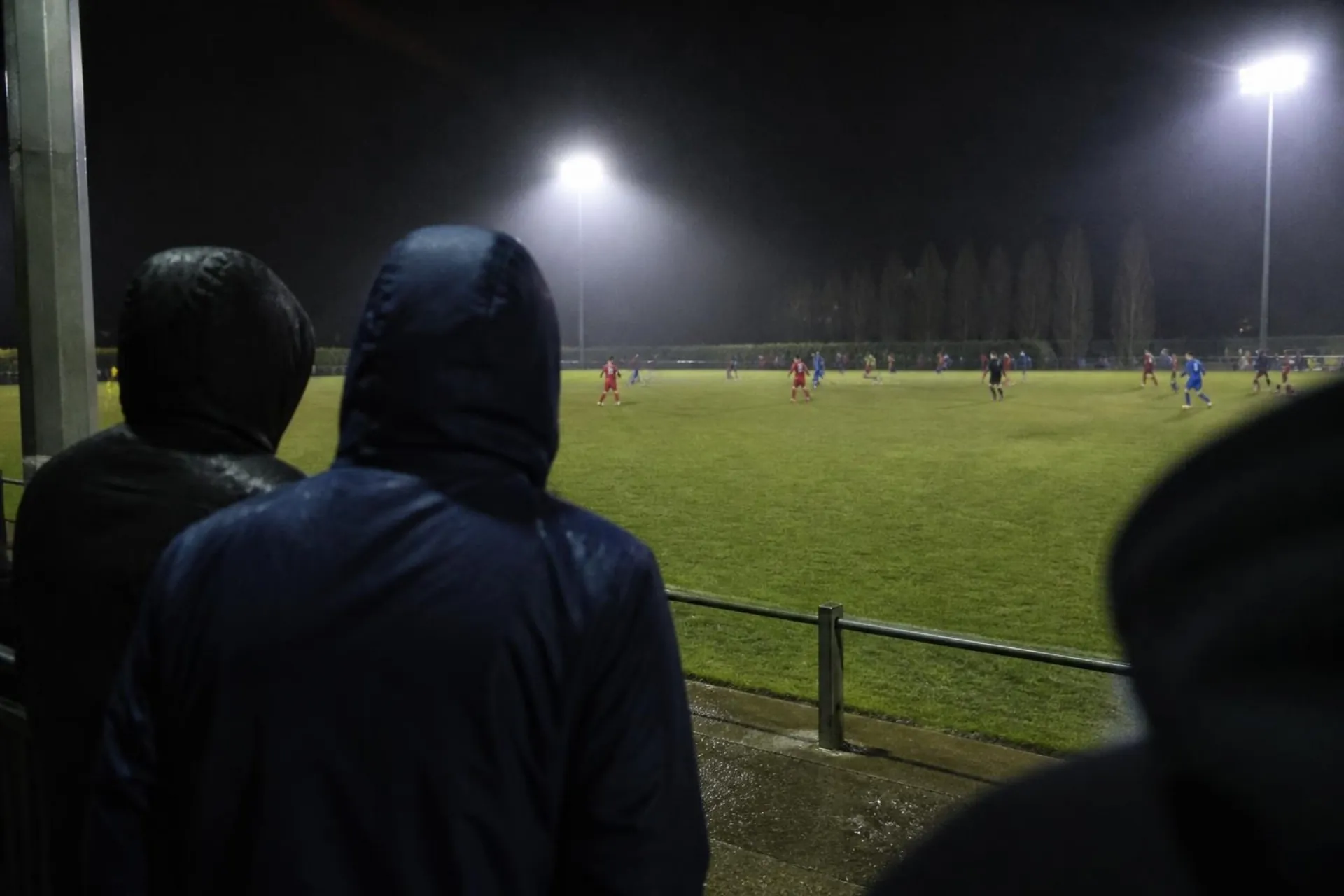 Hooded fans at a rail watching a floodlit lower-league match in the rain