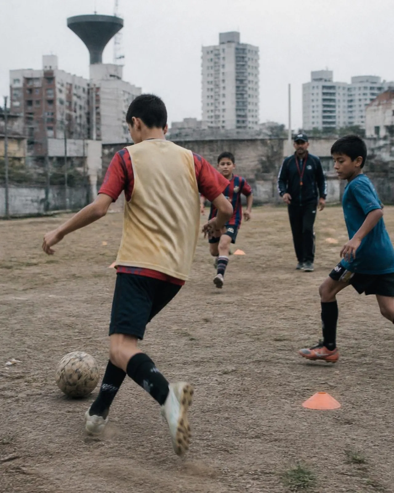 Kids in mixed kits running a dribbling drill on a dirt pitch, apartment blocks and a water tower behind